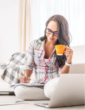 Woman having a tea, sitting on a floor in an apartment, writing down notes, opened laptop in front of her.